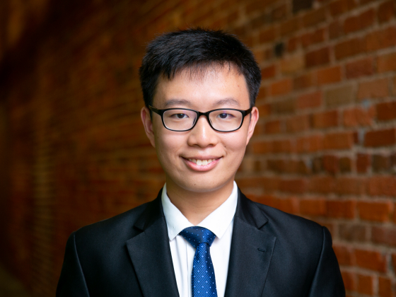 Asian man with glasses in a suit standing with a brick wall behind him