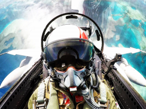A man in the cockpit of a fighter jet. There is water and earth in the background, he appears to be upside down. 