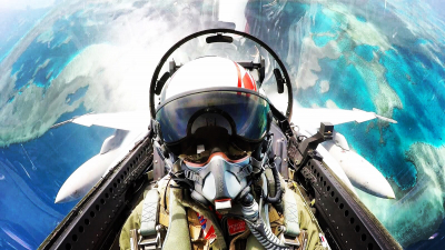 A man in the cockpit of a fighter jet. There is water and earth in the background, he appears to be upside down. 